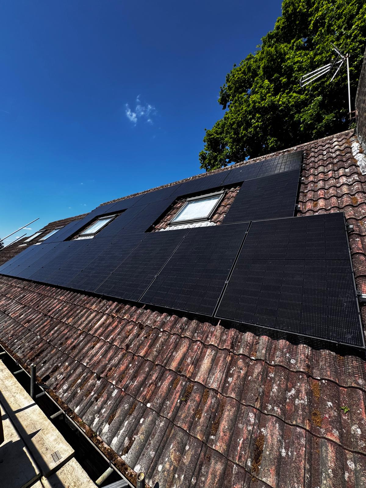Black solar panels fitted on a tiled roof around two skylights under a clear blue sky.