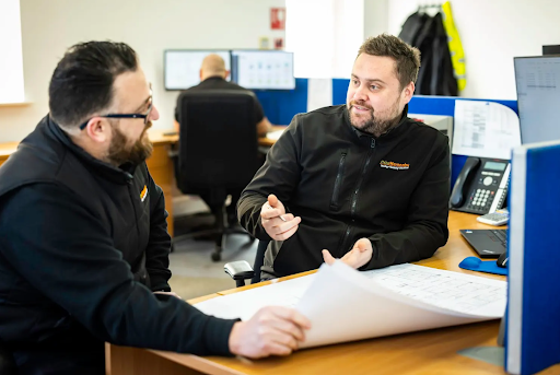 Two colleagues discuss technical drawings at a desk in an office, with another worker seated at a computer behind them.