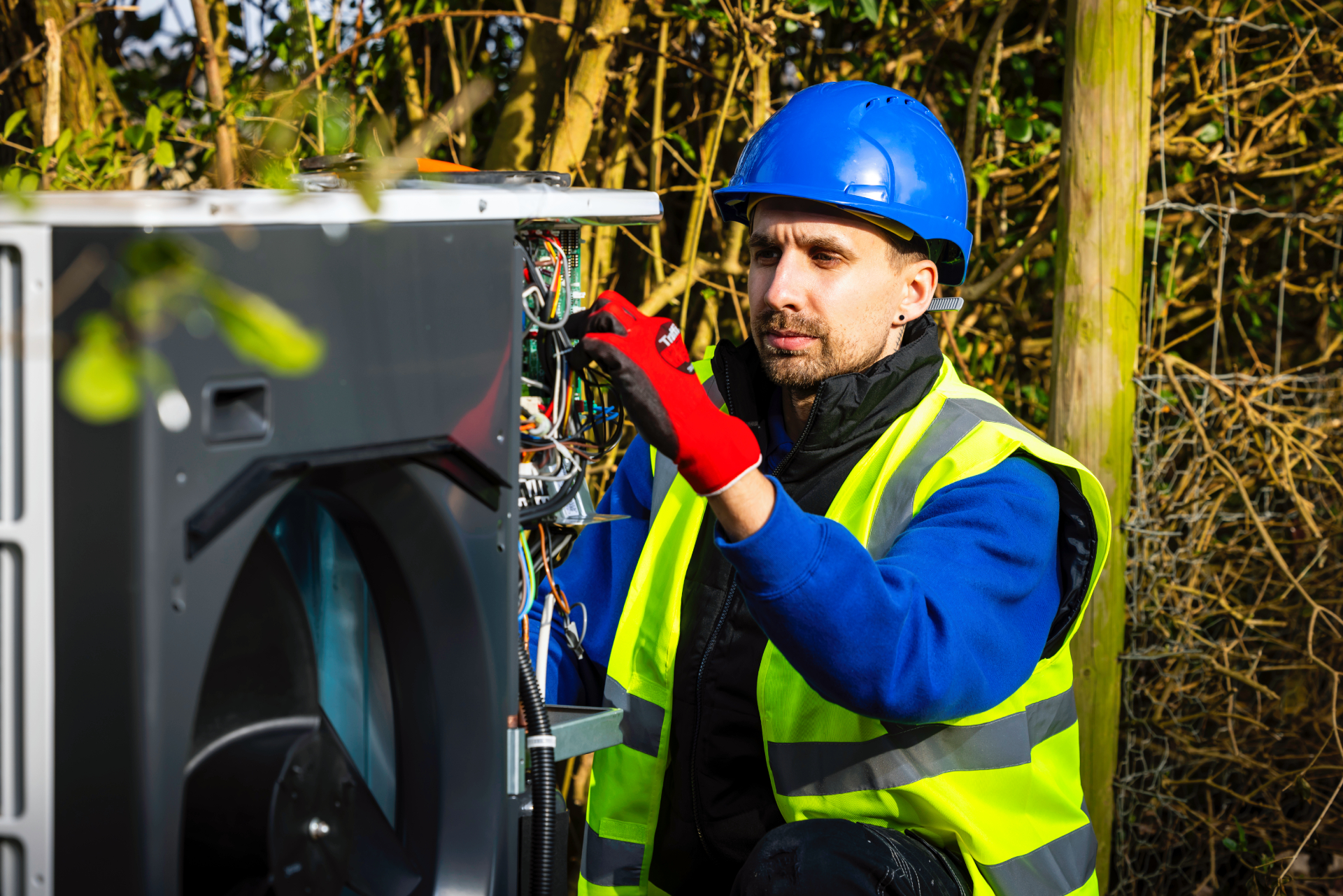 Engineer installing an air source heat pump outdoors while wearing safety gear, with the unit open and components visible. 
