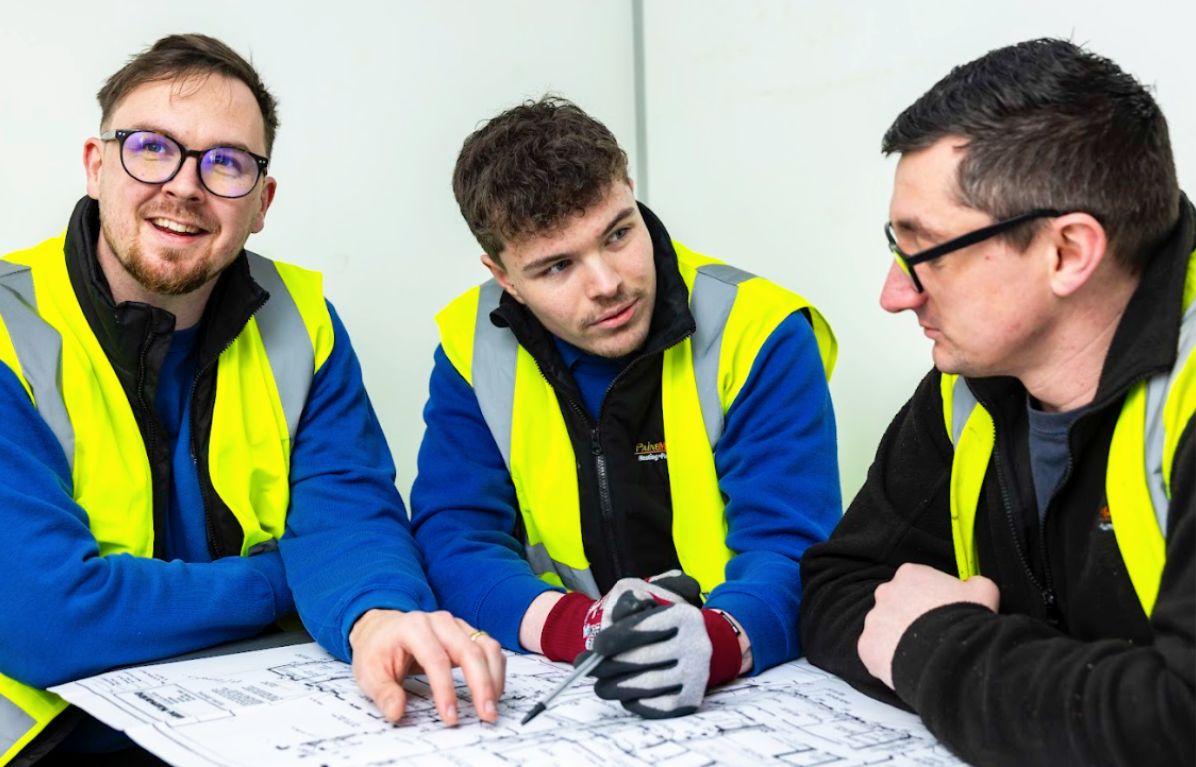 Three engineers reviewing technical drawings together while wearing high-visibility vests.